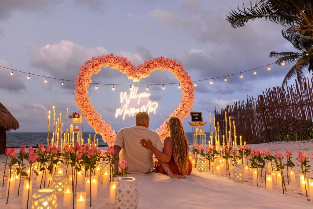 Luxury beach marriage proposal in Mexico with glowing heart-shaped floral arch and candlelit pathway at sunset, romantic beachfront engagement setup in Riviera Maya, elegant tropical proposal decor overlooking the Caribbean Sea, couple sitting together during intimate sunset proposal moment, warm golden-hour atmosphere with soft ocean breeze, sophisticated destination proposal styling in Mexico, cinematic beach romance with lanterns and flowers, premium luxury engagement experience, realistic tropical sunset lighting, highly photorealistic
