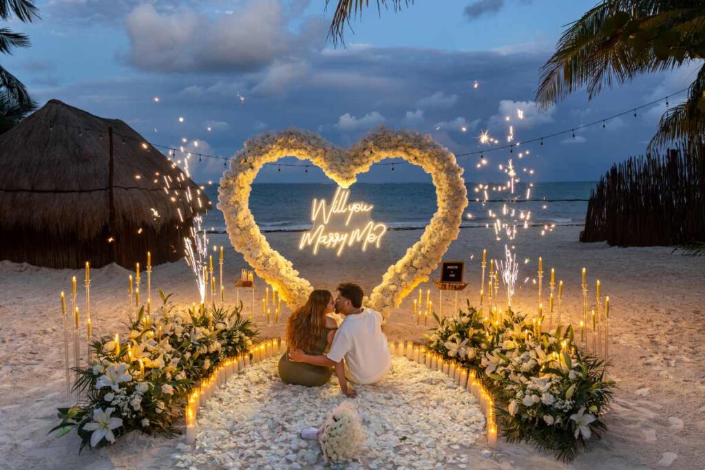 Luxury marriage proposal setup on the beach in Mexico with glowing heart-shaped lights, romantic sunset proposal decor overlooking the ocean, elegant candle pathway with white flowers, luxury destination engagement setup in Riviera Maya, cinematic beach proposal atmosphere, premium romantic experience in Mexico, boho luxury proposal styling at golden hour