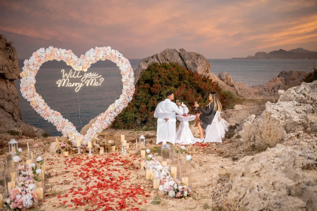 Luxury marriage proposal setup in Faliraki Rhodes overlooking the coastline at sunset, elegant heart-shaped floral arch with candlelit pathway on rocky cliffside, romantic beach proposal atmosphere in Rhodes Greece, sophisticated destination engagement decor with panoramic sea views, warm golden-hour light over the Aegean Sea, luxury sunset proposal styling with rose petals and lanterns, intimate Mediterranean proposal experience near Faliraki, cinematic coastal romance with realistic lighting and textures, premium proposal setup in Rhodes, highly photorealistic