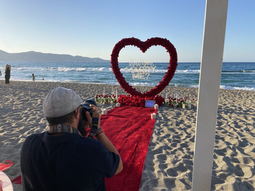 Luxury beach marriage proposal in Crete with red rose heart setup facing the sea, romantic proposal pathway on sandy beach in Greece, elegant candlelit engagement setup at sunset, luxury destination proposal in Crete, cinematic oceanfront proposal atmosphere, sophisticated romantic beach decor with red roses and lanterns, Mediterranean coastline in the background, emotional luxury proposal experience in Greece, premium sunset engagement setup, realistic and elegant proposal styling
